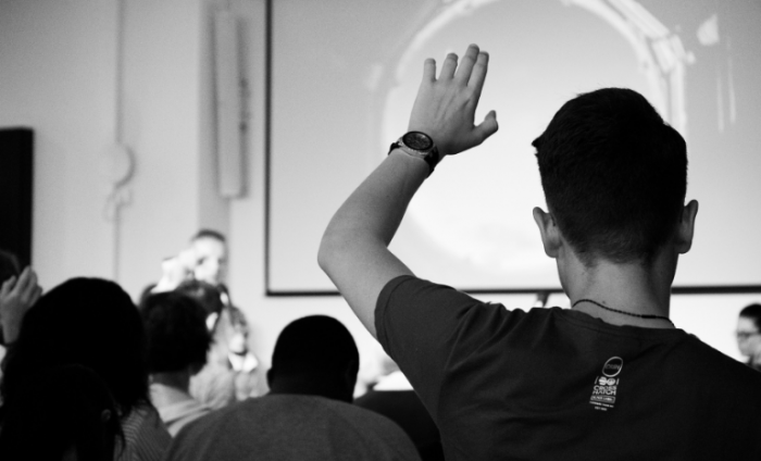 Black and white shot of an audience listening to a presentation and a man with his hand up to ask a question. 